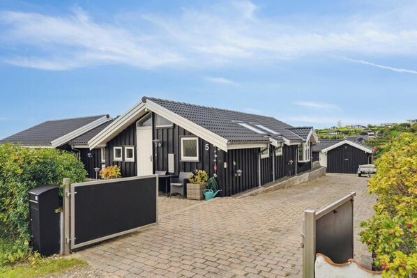 A row of modern, dark-colored cabins with a tiled roof, a paved driveway, and a fenced entrance. - Sea view holiday home near Ebeltoft (Ebeltoft)