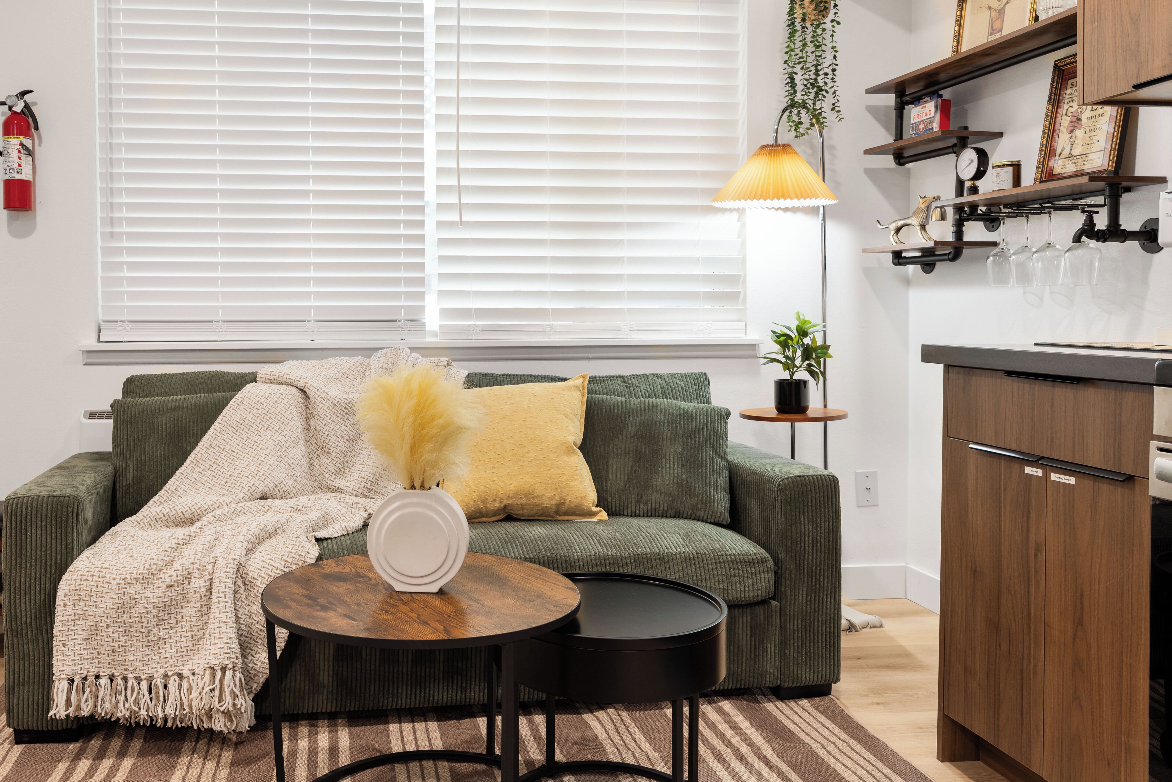 A living room with a sofa, a round coffee table, a lamp, and a kitchen area with wooden cabinets and shelves.