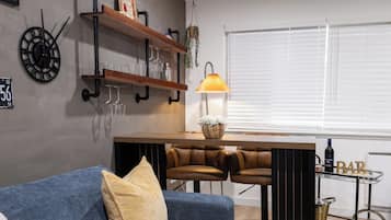 A modern living room with a blue sofa, two leather bar stools, a wooden coffee table, a wall-mounted clock, shelves with glassware, and a window with blinds.