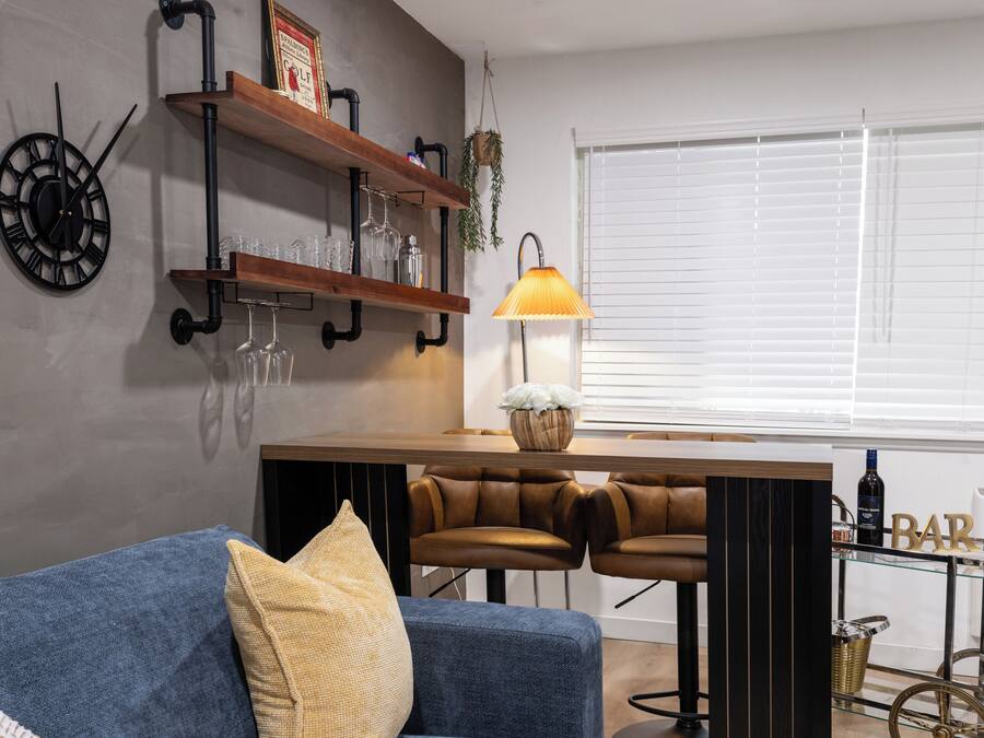 A modern living room with a blue sofa, two leather bar stools, a wooden coffee table, a wall-mounted clock, shelves with glassware, and a window with blinds.