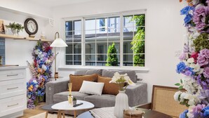 A living room with a grey sofa, a round coffee table, a white vase with flowers, and a large window with a view of greenery. - STAYOO SUITES-Seattle Secret Garden (Seattle)