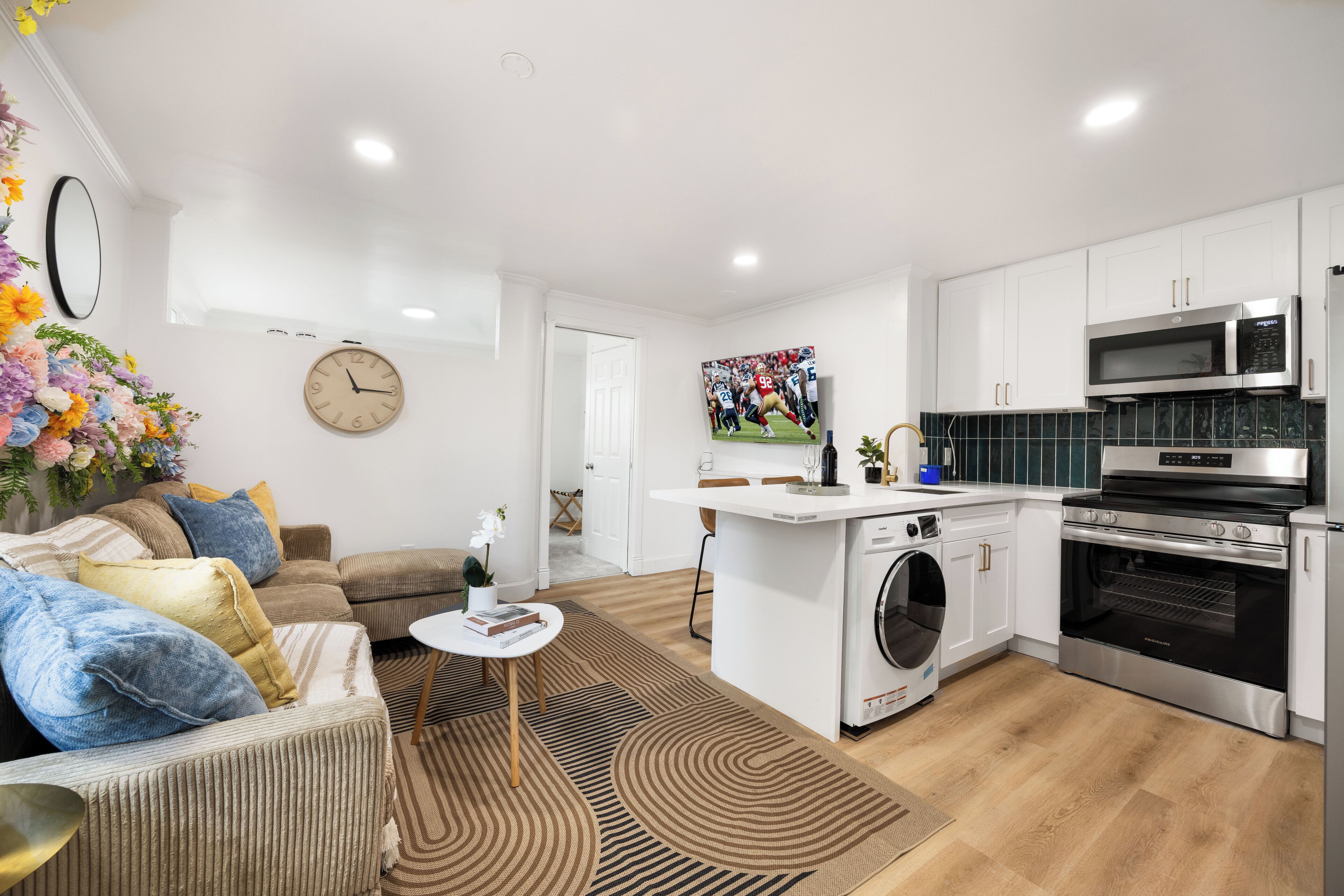 Een moderne keuken met een magnetron, oven en wasmachine. Er is een bank met kussens en een kleine tafel met een vaas.