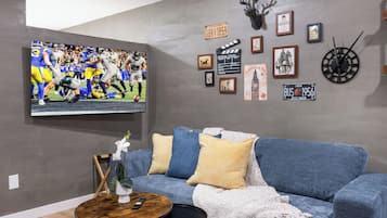 A living room with a blue sofa, a round coffee table, a mounted TV displaying a football game, and various framed pictures and a clock on the wall.