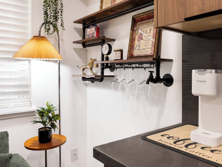 Una cocina con cafetera, una estantería con un certificado enmarcado, una planta colgante y una mesita con planta en maceta.