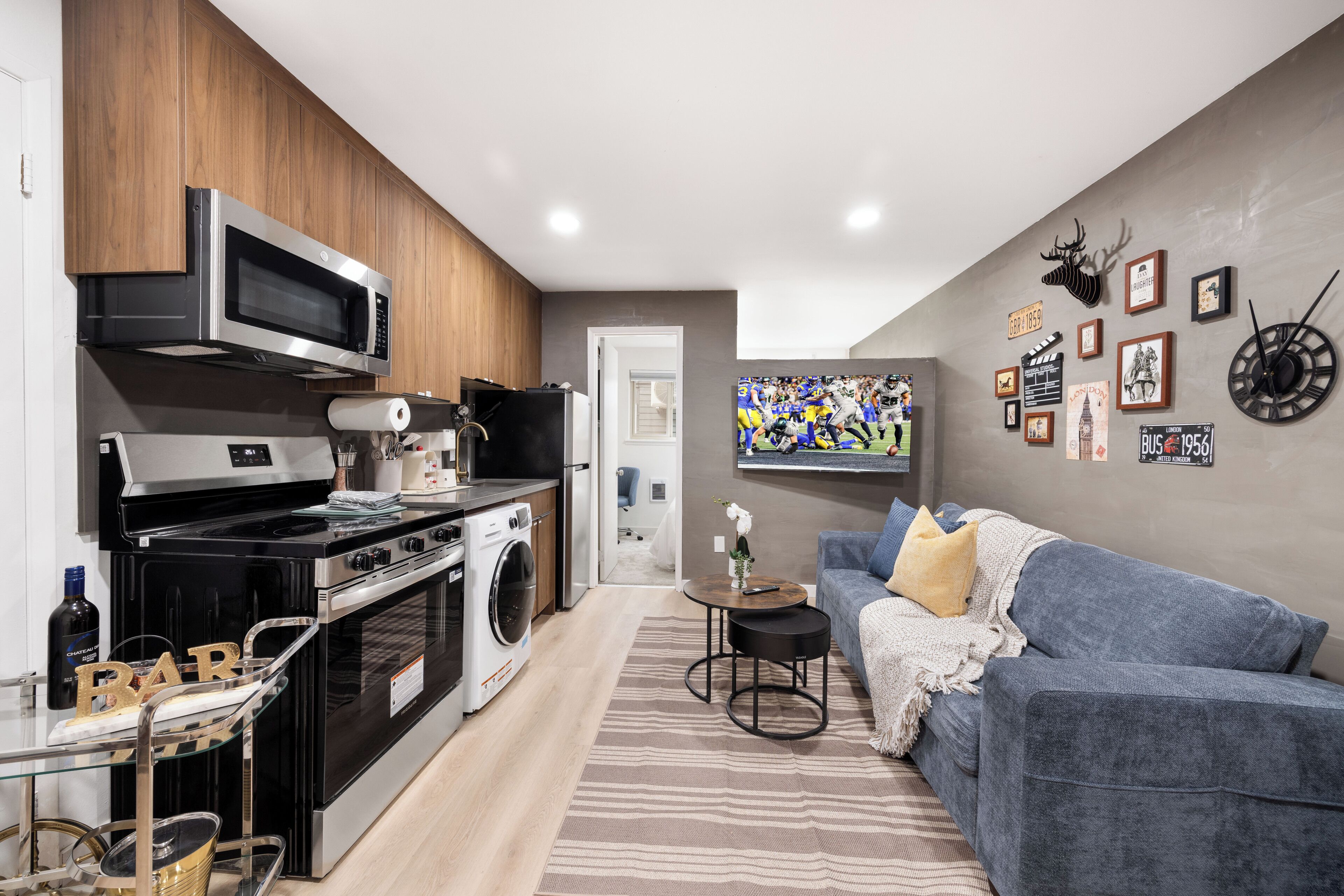 A modern kitchen with a microwave, stove, and washing machine, a gray sofa, and a wall with various framed pictures and a clock.