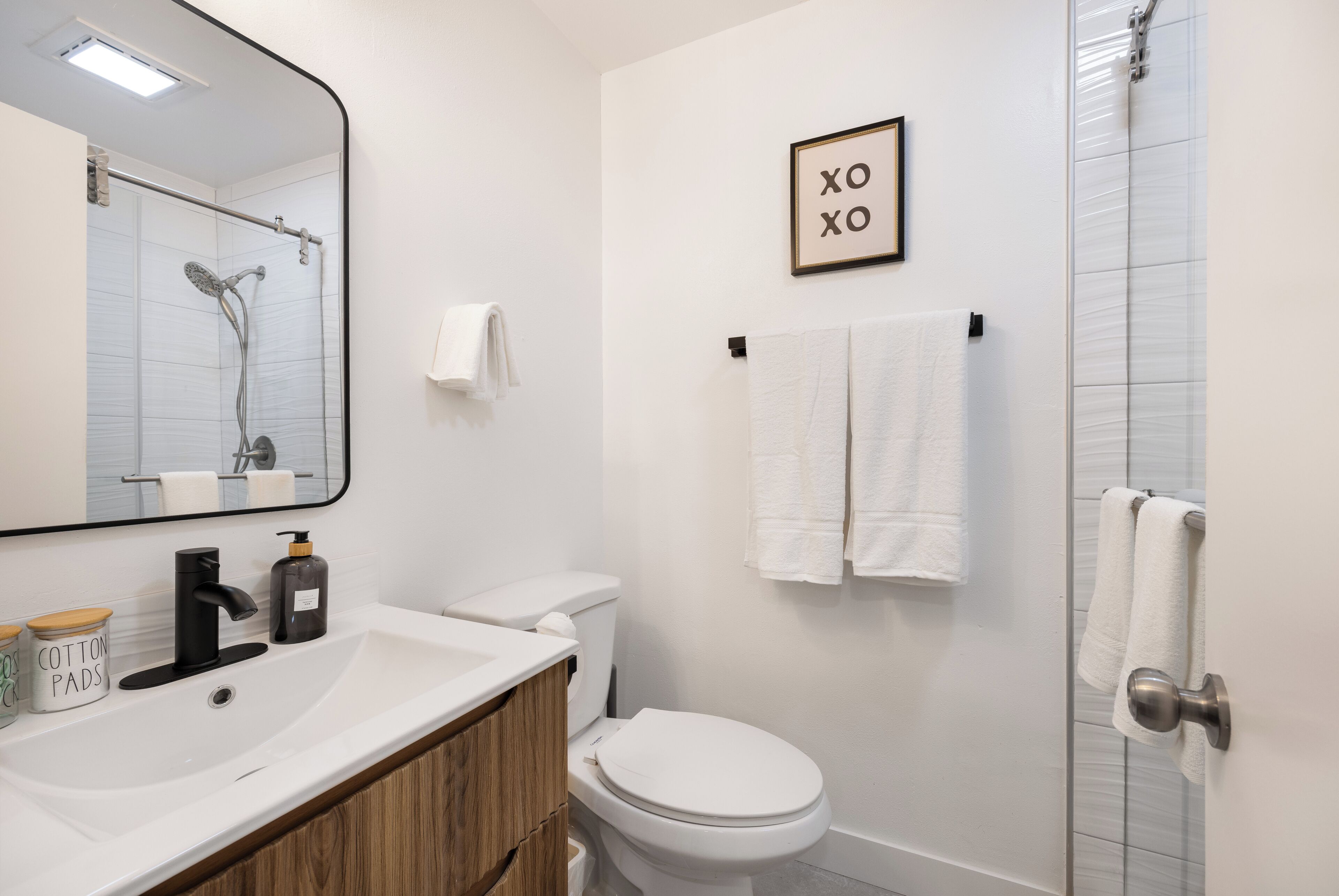 Un baño con lavamanos, inodoro y ducha blancos. Hay un cartel enmarcado en la pared y un toallero con toallas.