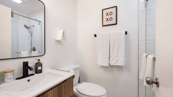 Un baño con lavamanos, inodoro y ducha blancos. Hay un cartel enmarcado en la pared y un toallero con toallas.
