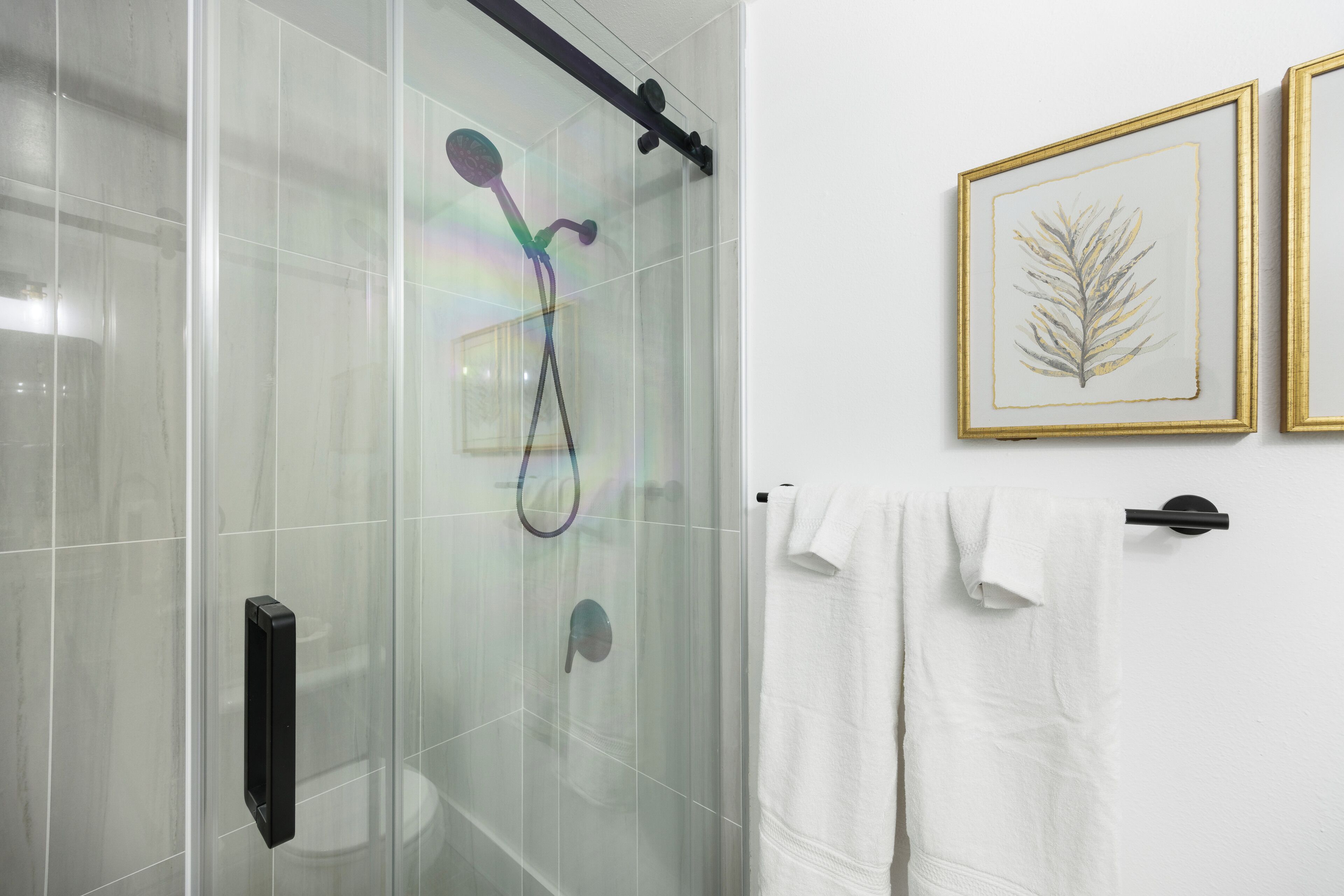Un baño con cabina de ducha de vidrio, un perchero con toallas blancas y dos obras de arte con hojas enmarcadas en la pared.