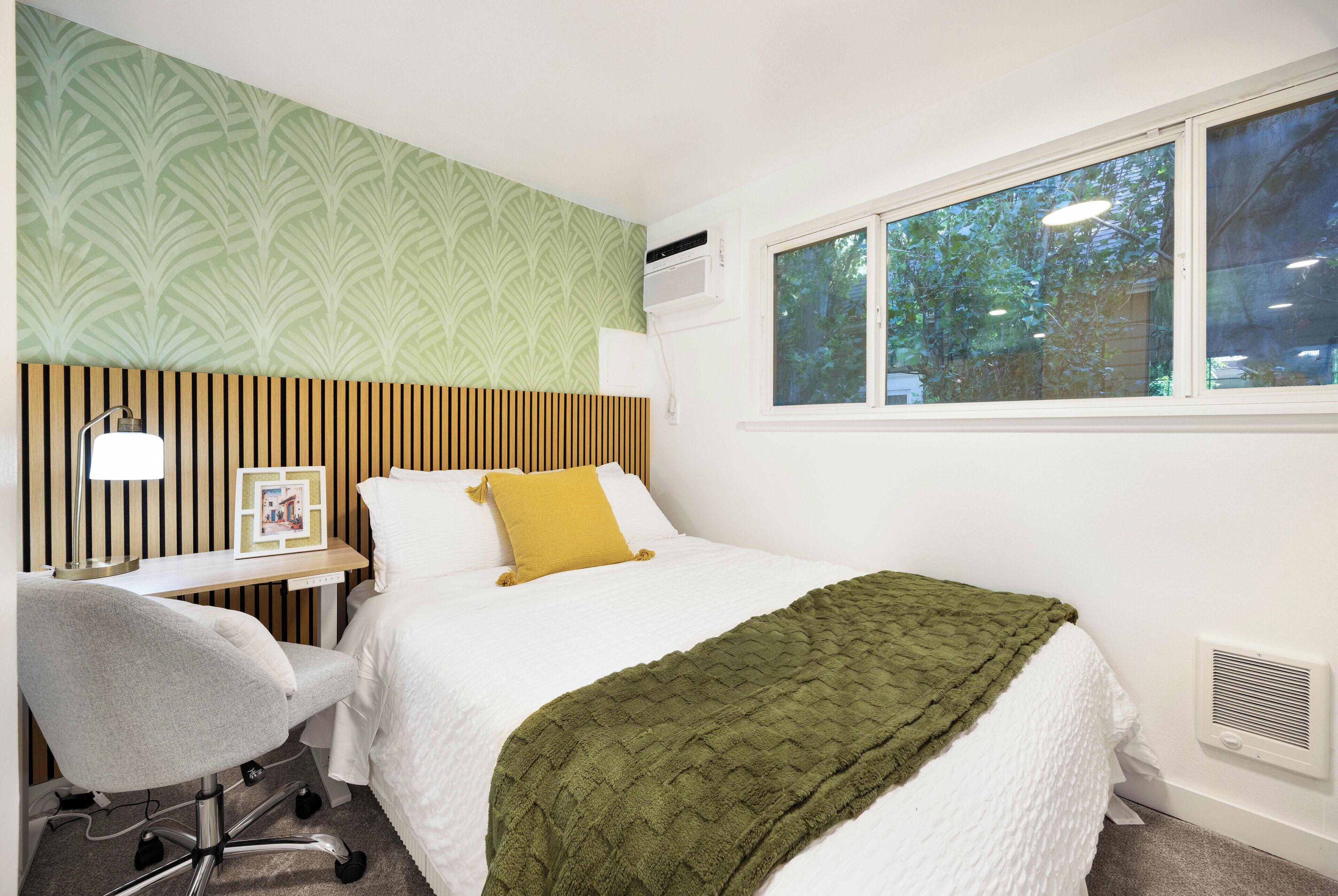 A hotel room with a bed, a desk with a chair, a window with a view of trees, and a wall with a patterned wallpaper.