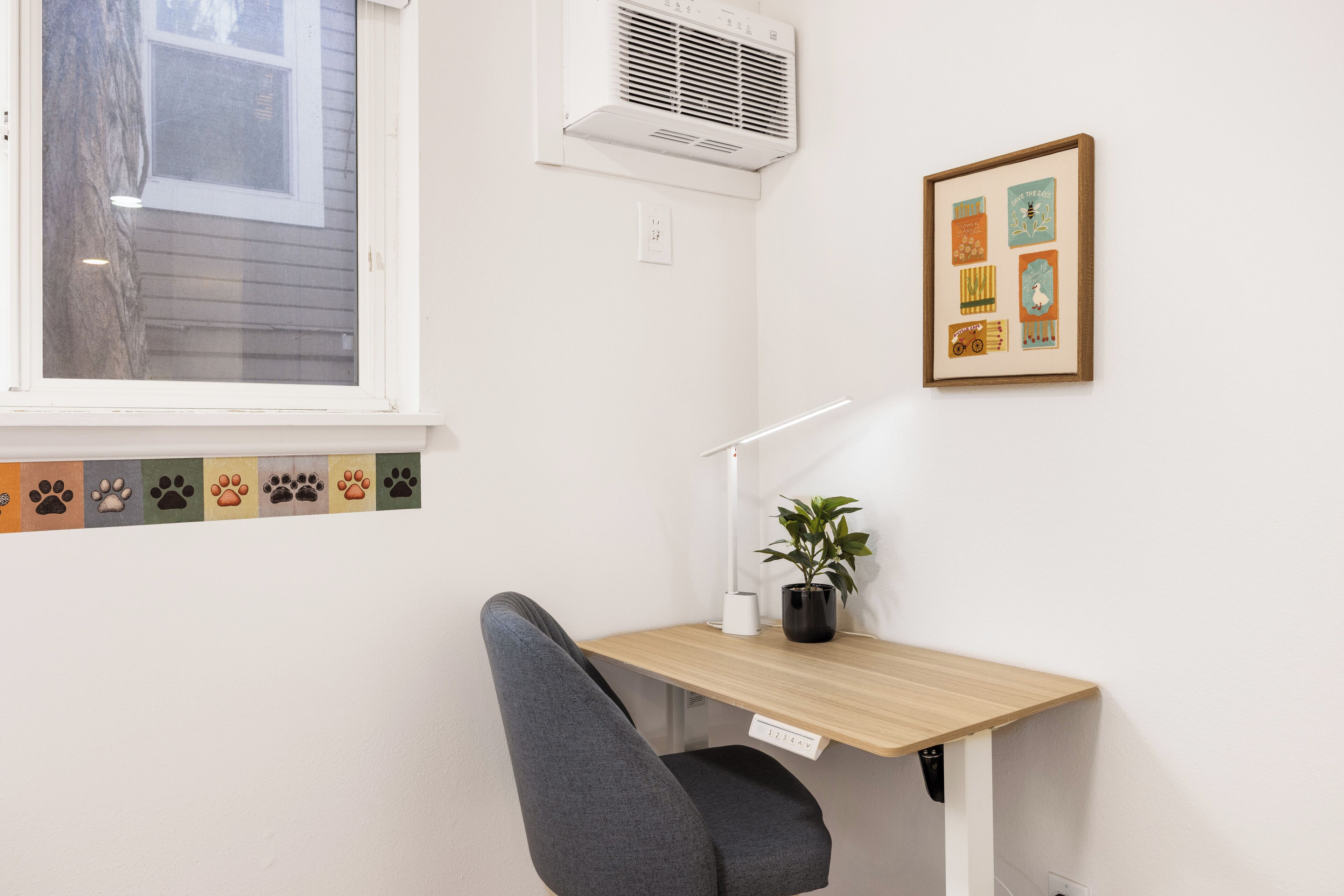 A room with a desk, chair, air conditioning unit, window, and a framed picture on the wall.