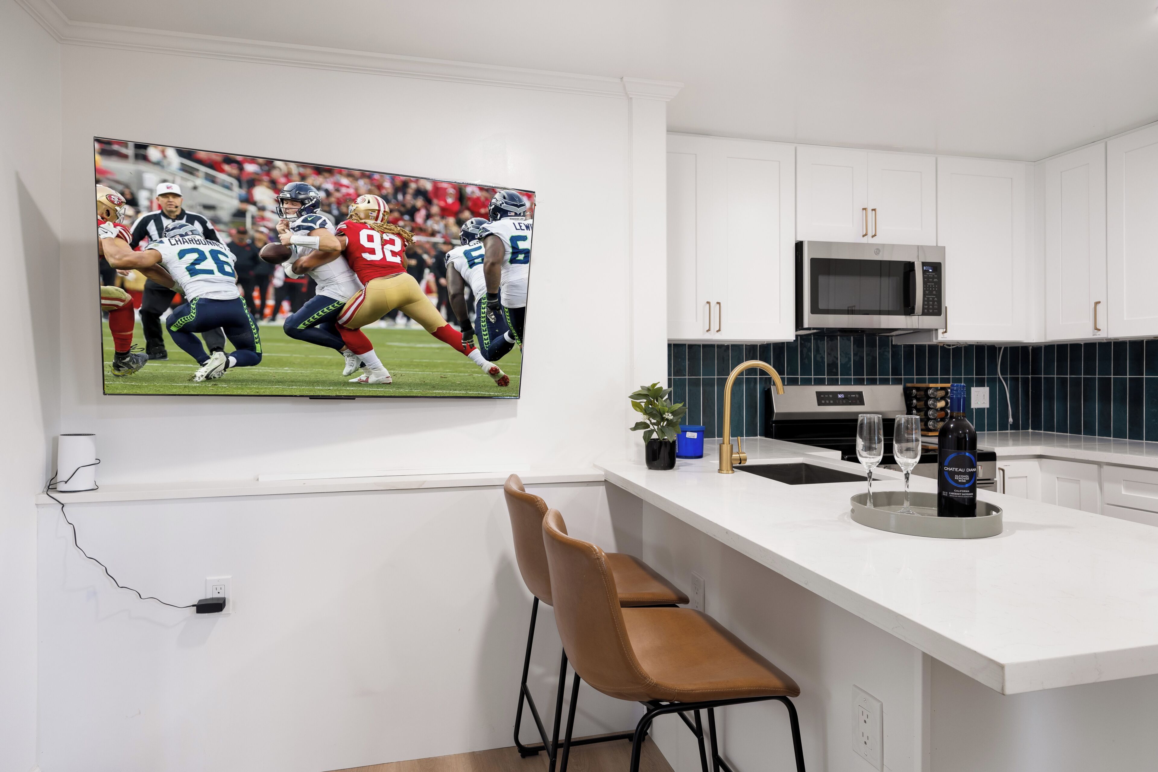 A kitchen with white cabinets, a stainless steel microwave, a bar area with a chair, and a TV displaying a football game.