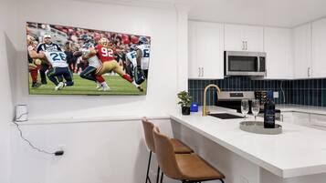 A kitchen with white cabinets, a stainless steel microwave, a bar area with a chair, and a TV displaying a football game.