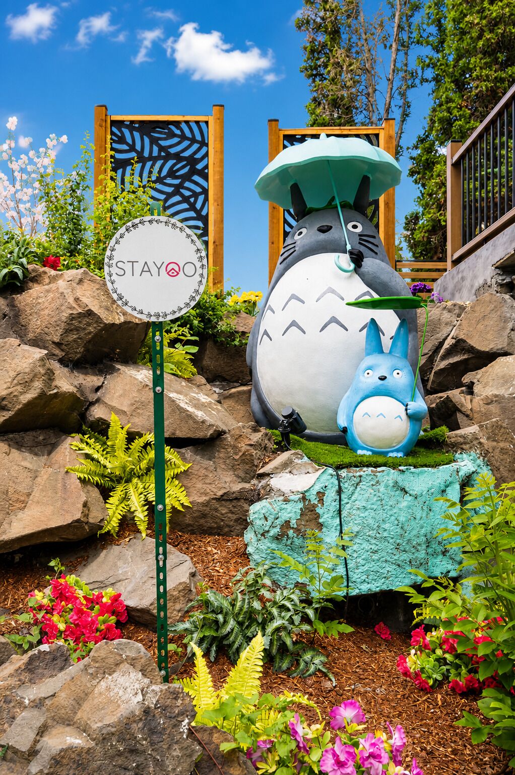 Una gran estatua de un personaje animado, un cartel con el logotipo 'STAYO', y un jardín con rocas y plantas.