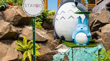 Una gran estatua de un personaje animado, un cartel con el logotipo 'STAYO', y un jardín con rocas y plantas.