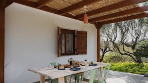 Un área de comedor al aire libre con una mesa y sillas de madera, un lámpara colgante, y vistas a árboles y follaje.