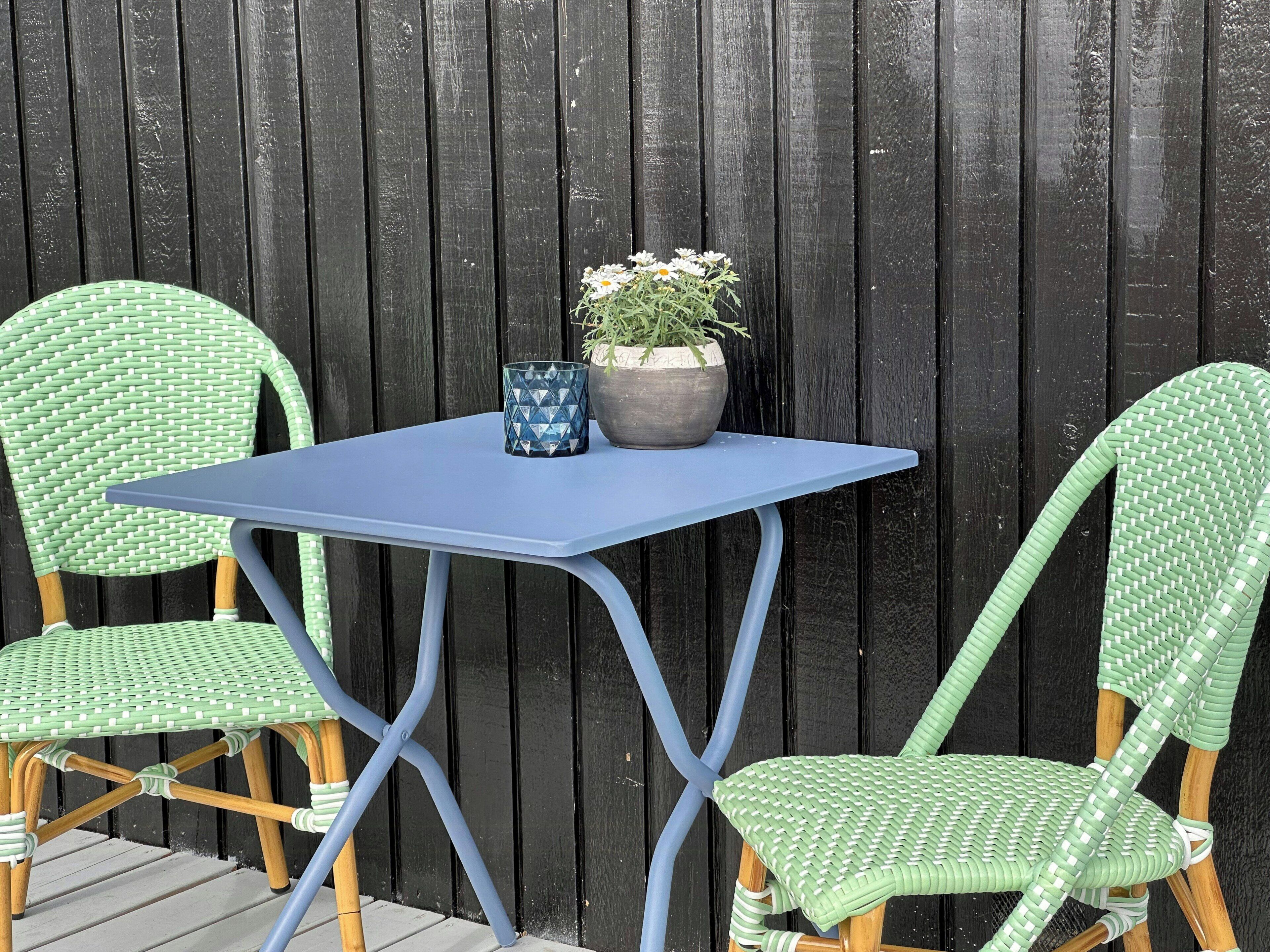 Une table bleue, deux chaises en osier vert et un pot de fleurs avec des fleurs blanches.