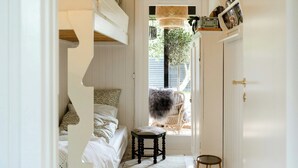 Quarto pequeno com uma cama, um banquinho, uma cadeira e uma porta para o exterior.