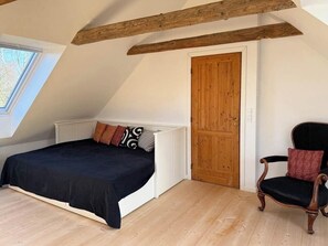A bedroom with a sloped ceiling, a bed with a black bedspread, a wooden door, and a chair with a cushion. - Renovated stone house from 1877 in Listed Bornholm (Svaneke)