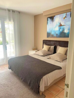 Ein Schlafzimmer mit einem großen Bett, einem Nachttisch, einem Bild an der Wand und Blick durch ein Fenster.