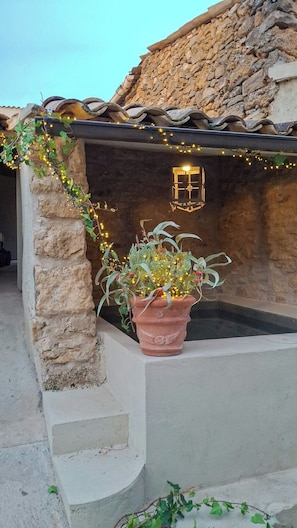 Un bâtiment en pierre avec un pot en terre cuite contenant des plantes, une petite fenêtre et des guirlandes lumineuses.