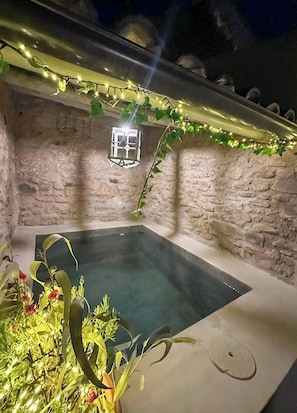 Un espace avec un bain à remous aux murs en pierre, doté d’une petite fenêtre, entouré de verdure et illuminé de guirlandes lumineuses.