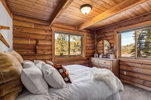 Ein Schlafzimmer in einer Blockhütte mit Bett, Kommode und großen Fenstern.