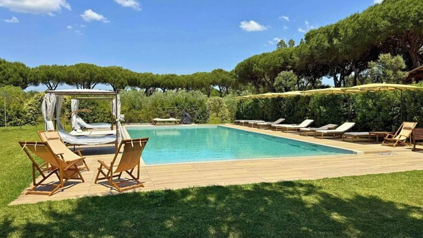 A rectangular swimming pool with a wooden deck, surrounded by lounge chairs and a canopy structure. - Private villa among pine trees and lavender (Marina Di Grosseto)