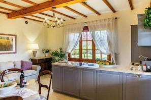 A kitchen with a central island, wooden beams, a chandelier, a window with curtains, and a dining area with a sofa and table.