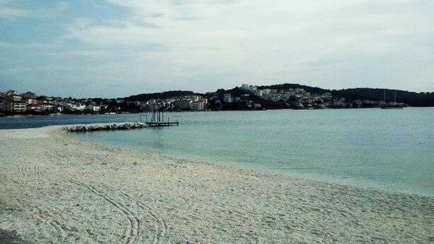 En strand med klart vand, en moler og en fjerbelliggende bakke med bygninger.