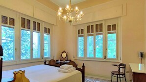 Una habitació amb un llit, una cadira, un escriptori, una làmpada de cristall i finestres grans amb persianes.