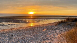 Ein Strandbild mit untergehender Sonne über dem Wasser, einem sandigen Pfad, der zum Wasser führt, und etwas Gras im Vordergrund.
