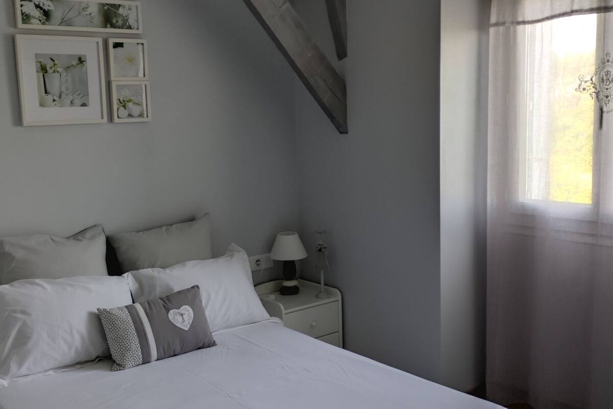 Un dormitorio con cama, dos mesitas de noche con lámparas, una ventana con cortinas y cuadros en la pared.