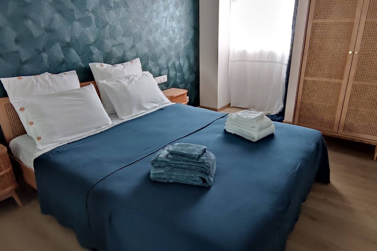 Una cama bien hecha con almohadas blancas y una colcha azul, una mesita de noche de madera, una cesta de mimbre y una pila de toallas dobladas.