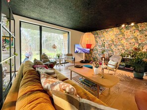 A modern living room with a large sofa, a coffee table, and a TV. There is a large window providing a view of the outdoors. - Holiday home with forest view 200 m from beach (Tved)