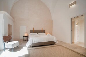 Ein Schlafzimmer mit einer hohen, gewölbten Decke, einem Bett mit weißen Bettwäsche, einem Holz-Kopfteil, einem kleinen Nachttisch mit Lampe und einem Sessel.