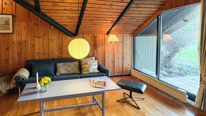 Een gezellige kamer met een houten plafond, een bank met kussens, een tafel met een bloevas en een raam met uitzicht op bomen.