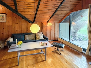 Ein gemütliches Zimmer mit Holzbalkendecke, einer Couch mit Kissen, einem Tisch mit Blumenvase und einem Fenster mit Blick auf Bäume.