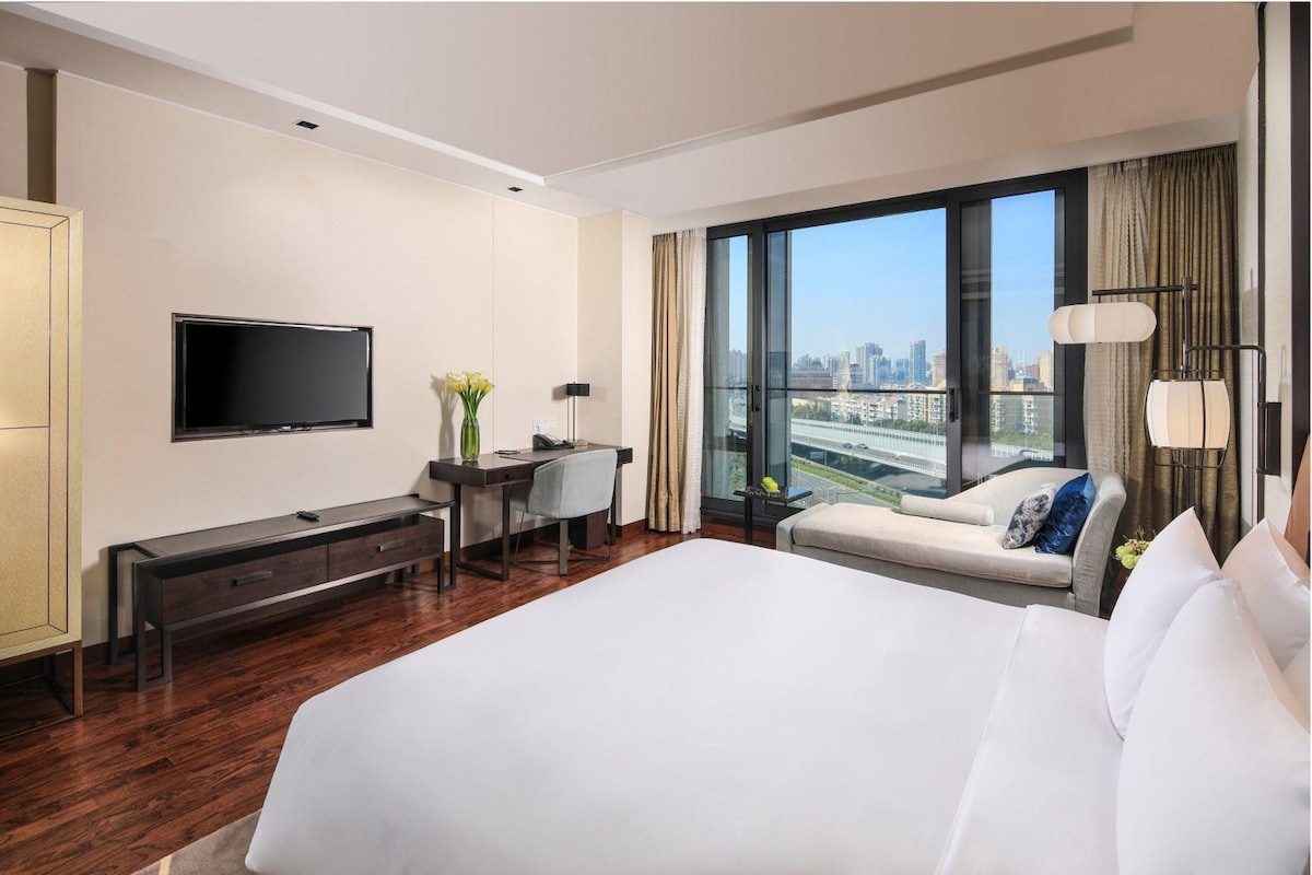 Habitación de hotel con una cama grande, un escritorio con silla, televisión y vista a un paisaje urbano a través de amplias ventanas.