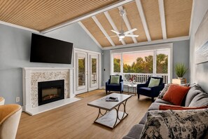 A living room with a fireplace, a television, a ceiling fan, and a view of the outdoors. - Shared Pool + Pickleball Court + Sunroom | Walk to the Beach | Isle Be There (Isle Of Palms)