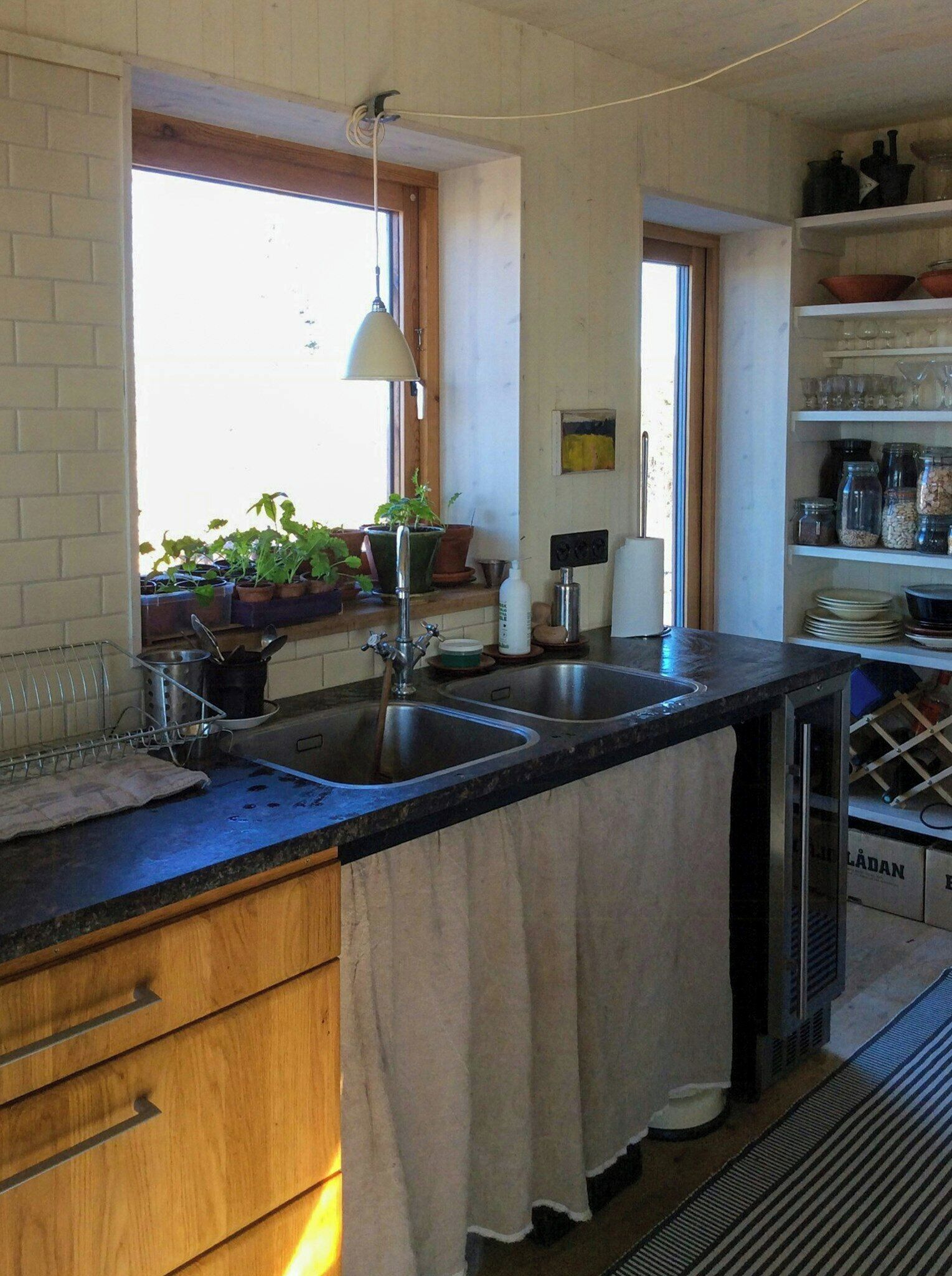 A kitchen with a double sink, wooden cabinets, a window with plants, and a hanging light fixture.