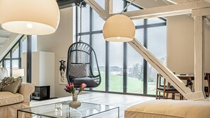 Un loft spacieux avec de hauts plafonds, de grandes fenêtres, un fauteuil suspendu, une table basse en verre et un coin salon confortable.