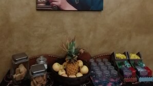 A wall with a framed picture of a person drinking from a cup, a fruit bowl with pineapple and apples, and various containers of snacks.
