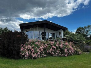 Ein modernes Haus mit einem großen Glasfenster, umgeben von üppigem Grün und rosafarbenen Blüten.