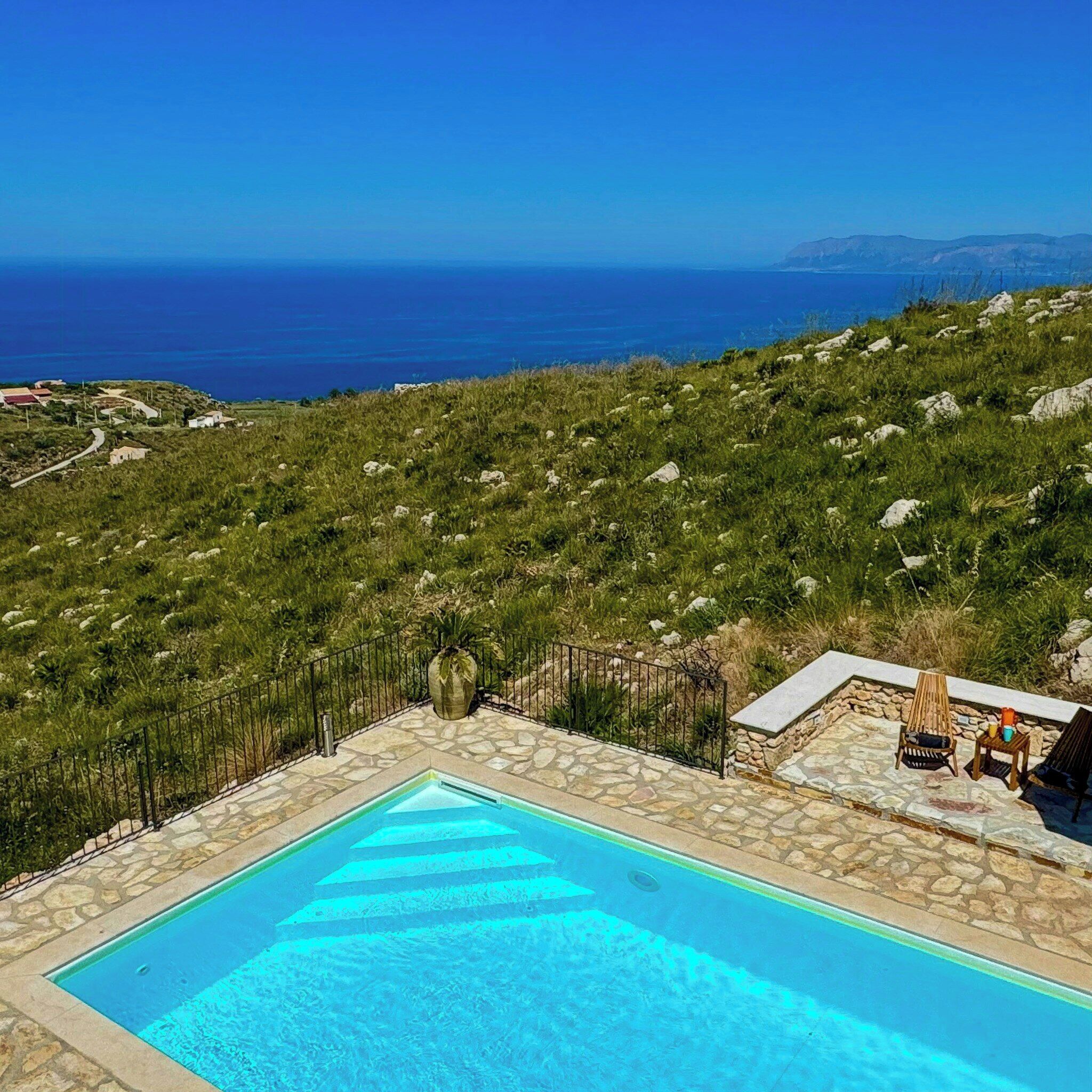 A pool with a clear view of the sea, surrounded by a stone deck and a grassy hillside.