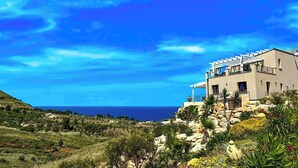 A modern two-story building with a balcony, surrounded by rocky terrain and vegetation, overlooking a coastal area. - Sea view villa with panoramic pool near Scopello (Castellammare del Golfo)