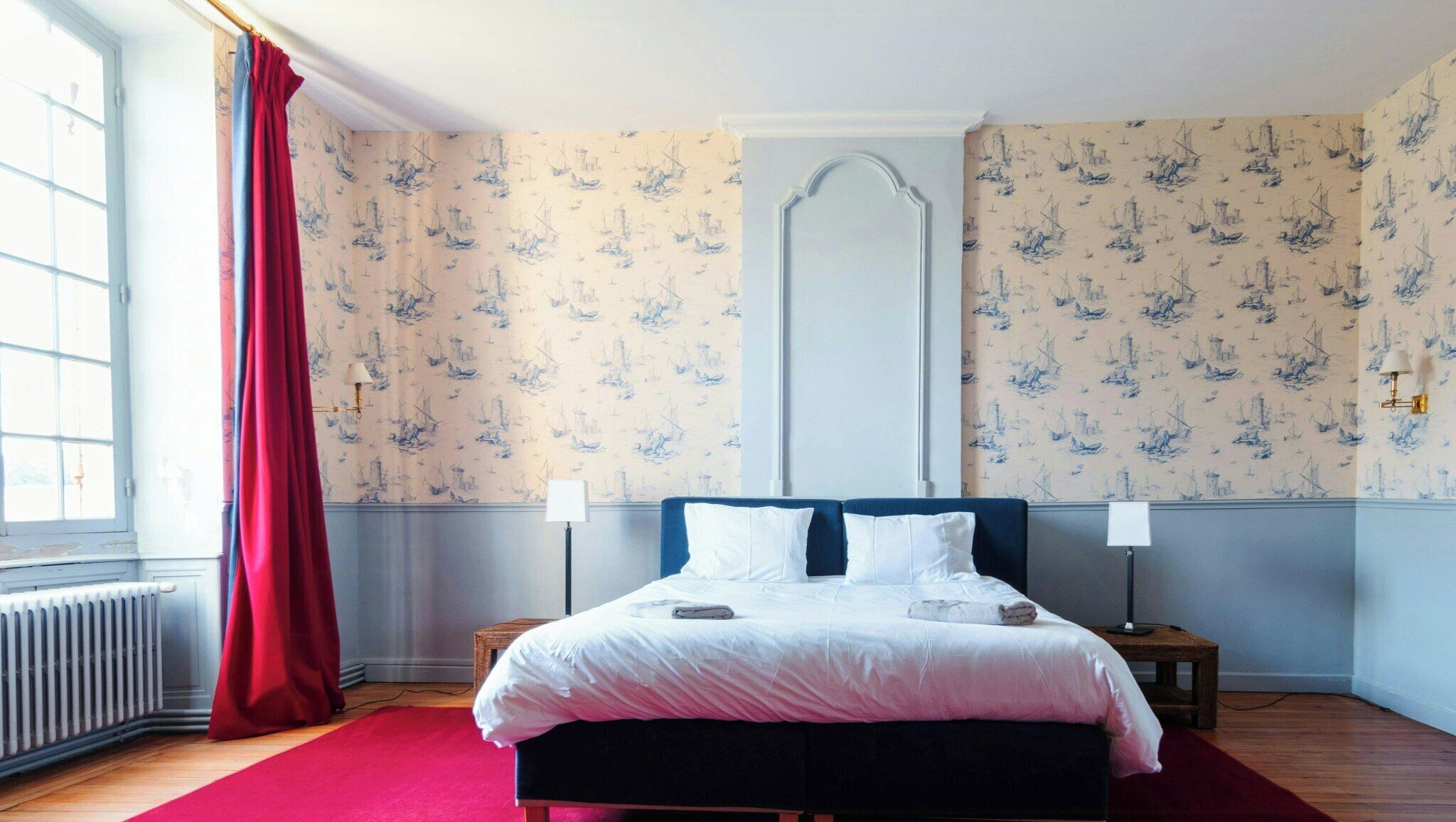 A bedroom with a bed, two bedside tables, a radiator, a window with a red curtain, and a wall with a nautical-themed wallpaper.