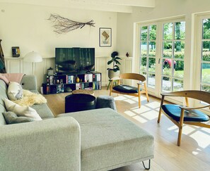 Ein Wohnzimmer mit Sofa, Couchtisch, Fernseher, Bücherregal und einem großen Fenster mit Blick auf Grünflächen.