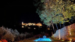 A nighttime view of a pool area with lounge chairs, a well-lit building in the background, and a stone wall with lights. - Bohemian house overlooking Roussillon (ROUSSILLON)