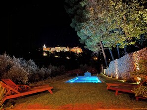 A nighttime view of a pool area with lounge chairs, a well-lit building in the background, and a stone wall with lights. - Bohemian house overlooking Roussillon (ROUSSILLON)