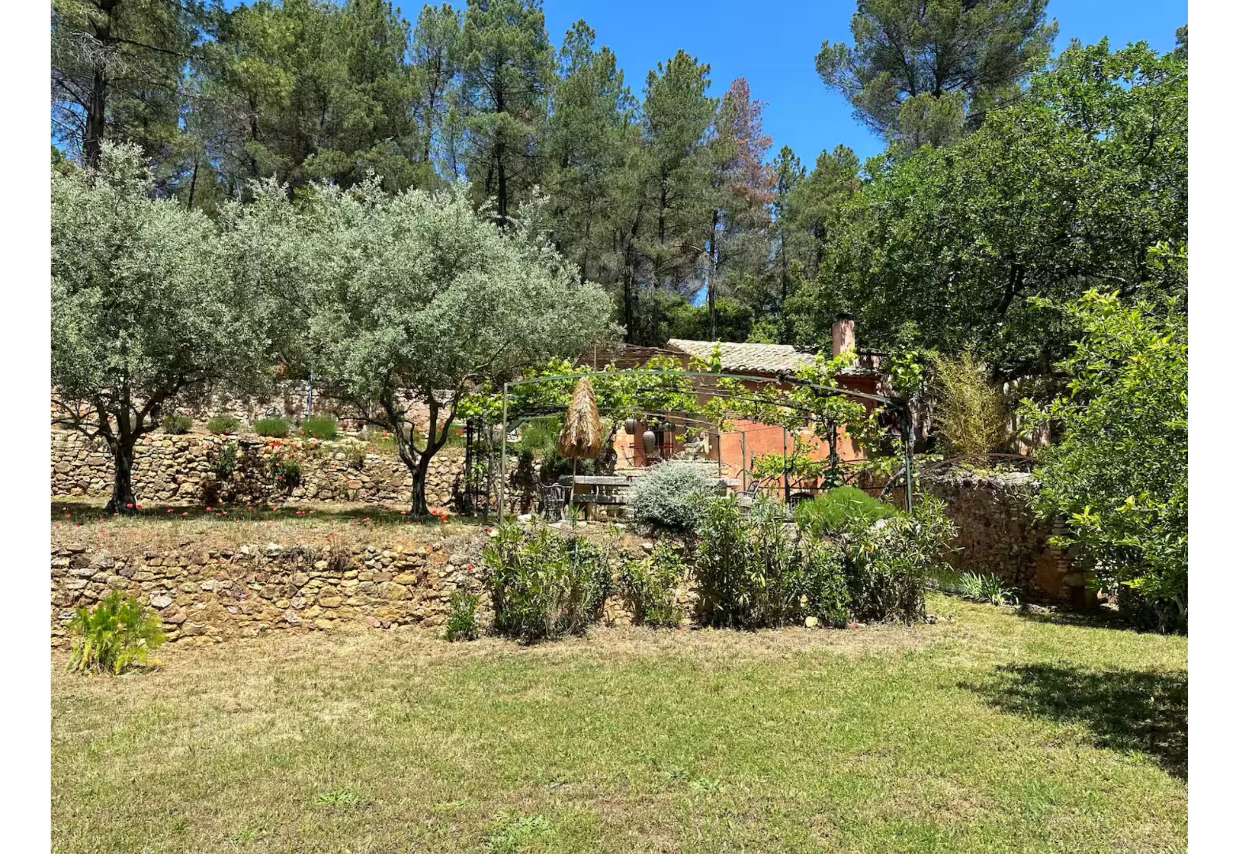 Un bâtiment rustique en pierre, doté d’un toit en tuiles, entouré d’un jardin bien entretenu où poussent différentes espèces d’arbustes et d’arbres.